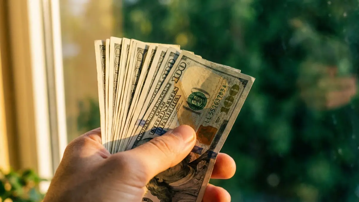 a hand holding a spread of US dollar bills with golden morning light streaming across the scene, representing daily money affirmation practice