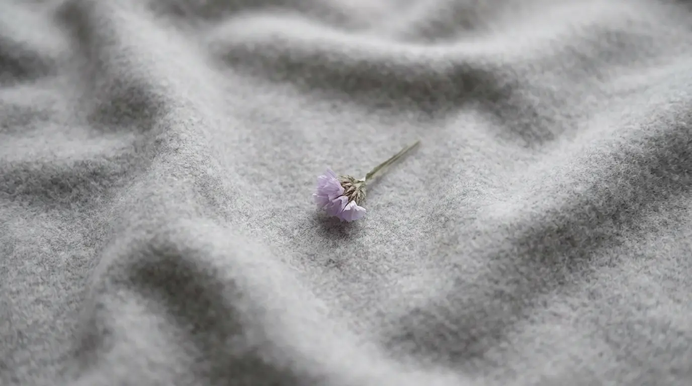 A flat lay of soft grey wool fabric filling the entire frame with natural gentle creases, one small pale lavender dried flower placed slightly off-center on the grey surface, overcast diffused cold light from above creating almost no shadows, photorealistic textile photography, muted grey-pearl tones with the single lavender accent as the only color in the frame, shallow depth of field with the flower in sharp focus, no text in the image, 16:9 ratio. Maintain visual consistency: grey-pearl and graphite palette, overcast diffused lighting, photorealistic textile style.