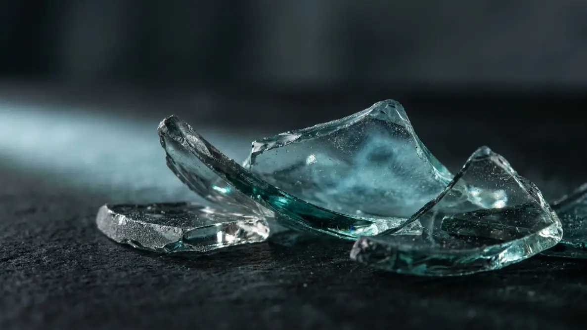 close-up of translucent glass fragments on a dark surface reflecting teal and silver light, representing the deep spiritual layers behind glass breaking without explanation