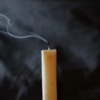 A candle with a smoking wick moments after the flame stopped, against a dark background