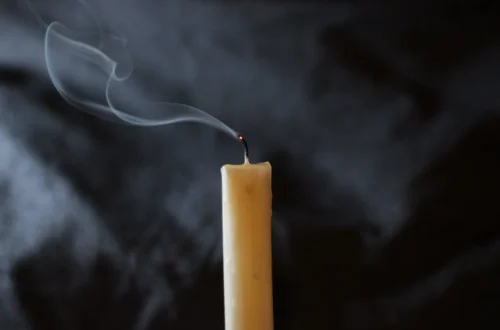 A candle with a smoking wick moments after the flame stopped, against a dark background