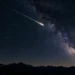A bright shooting star crossing the Milky Way above a dark mountain landscape.